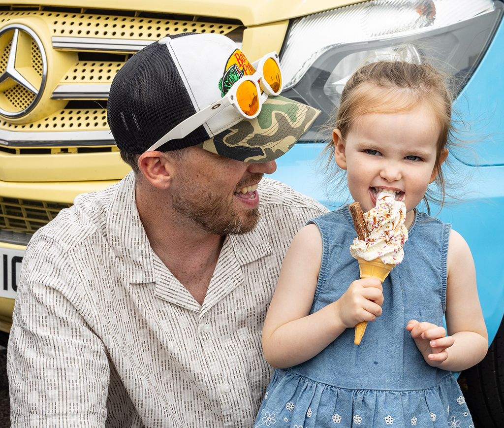 A smiling man wearing a camouflage peaked mesh baseball cap and sunglasses looks on as a young girl in a denim dress eats a soft-serve ice cream cone with sprinkles and a chocolate flake, with a yellow Mercedes-Benz vehicle in the background.
